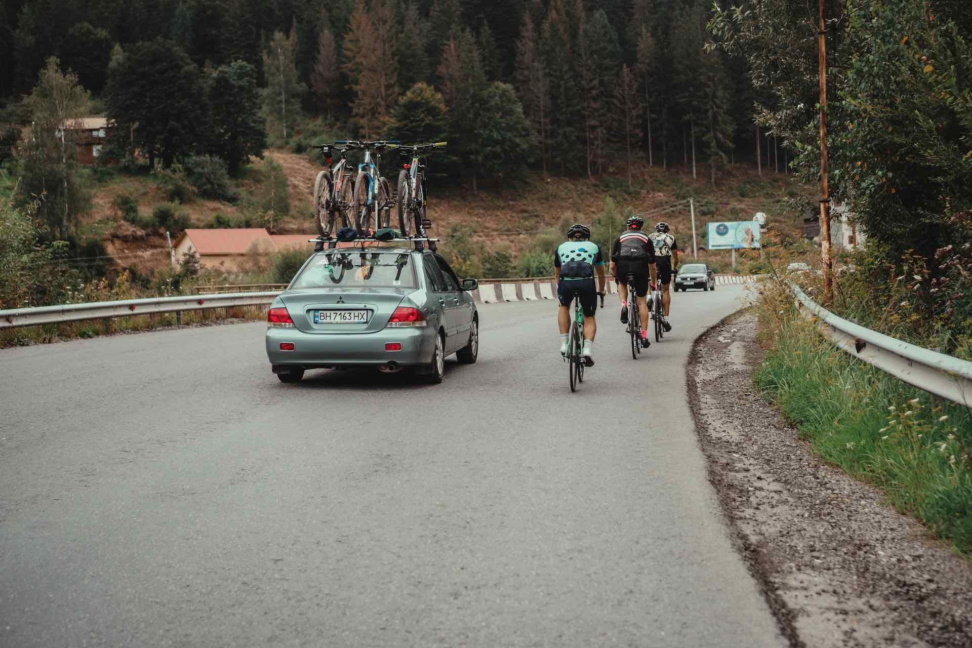 car with a cycling rack and 3 cyclists next to car on a mountain road