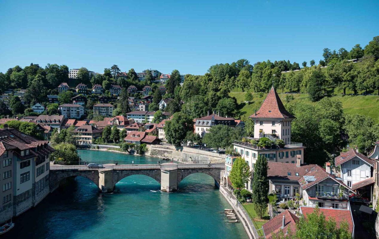 bern Switzerland a town with a river running through it with a road bridge across the river. the town is surrounded by green trees