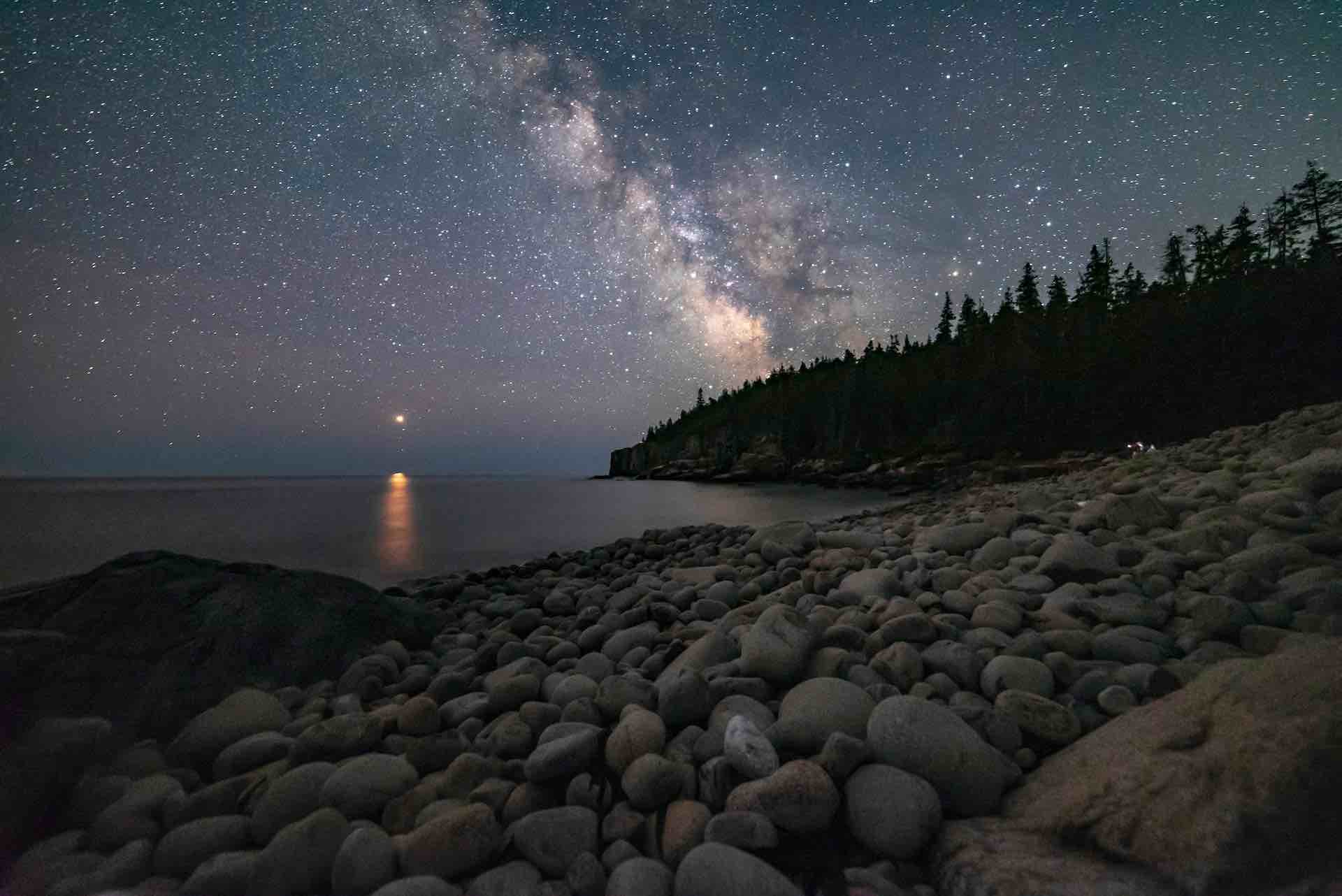 the nighttime view of Bar harbour in the Acadia National Park. Calm water with a silhouette of pine trees along shore. In the sky are twinkling stars, the moon is hidden behind the tree line but still lights the sky. In the distance over the water is a lone lit lighthouse. The sky is completely clear and the view to the horizon seems endless.