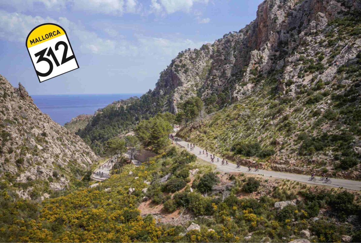 a mountain scene of the Mallorca 312 event, in the top left there I blue sky, the rest is barren mountains with some greenery. there is a road following the contour of the mountains and you can see the cyclists racing down hill on that road